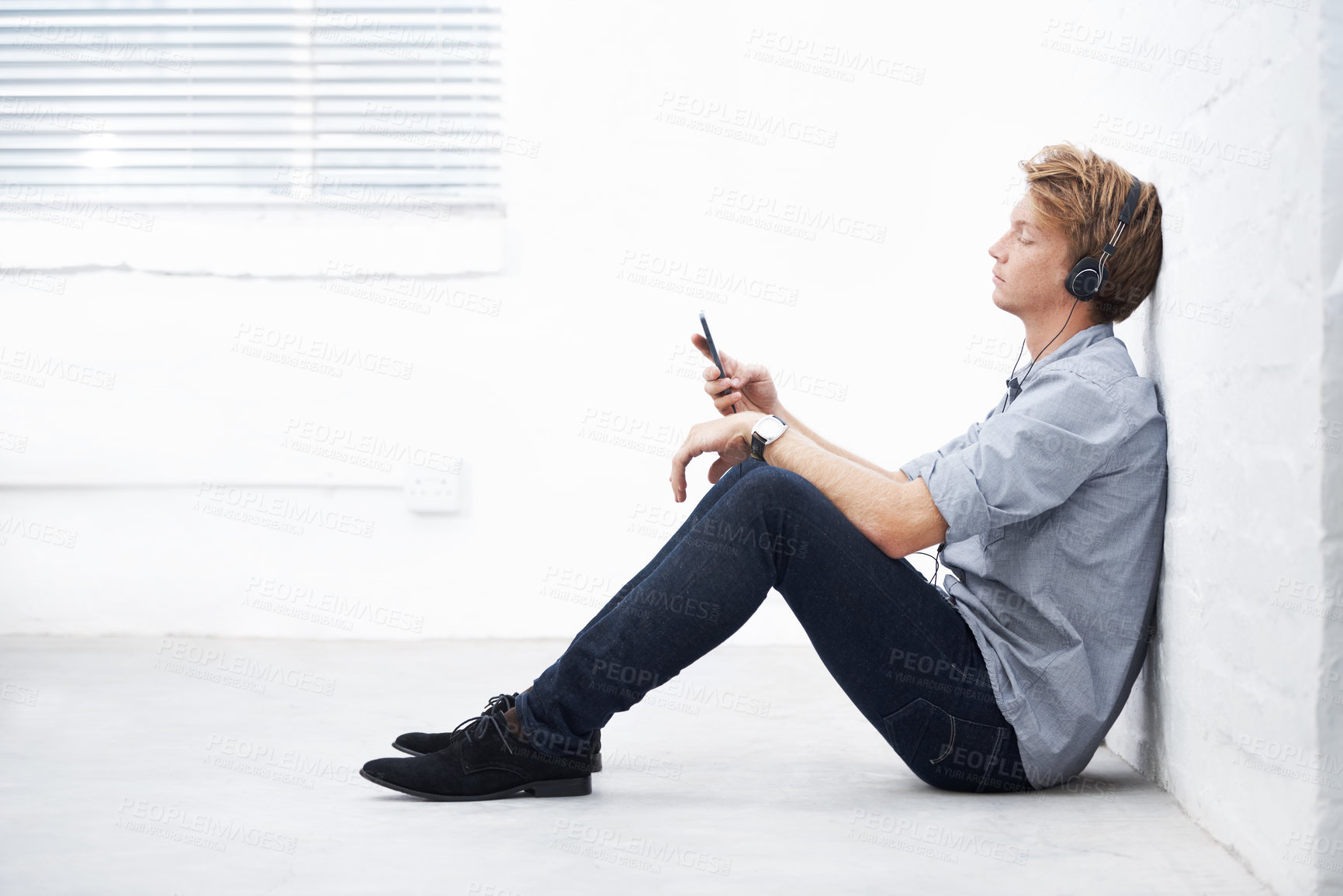 Buy stock photo Bored, tired designer relaxing, meditating, searching podcast during office break. Creative businessman listening to playlist music on phone with headphones, sitting against white wall with copyspace