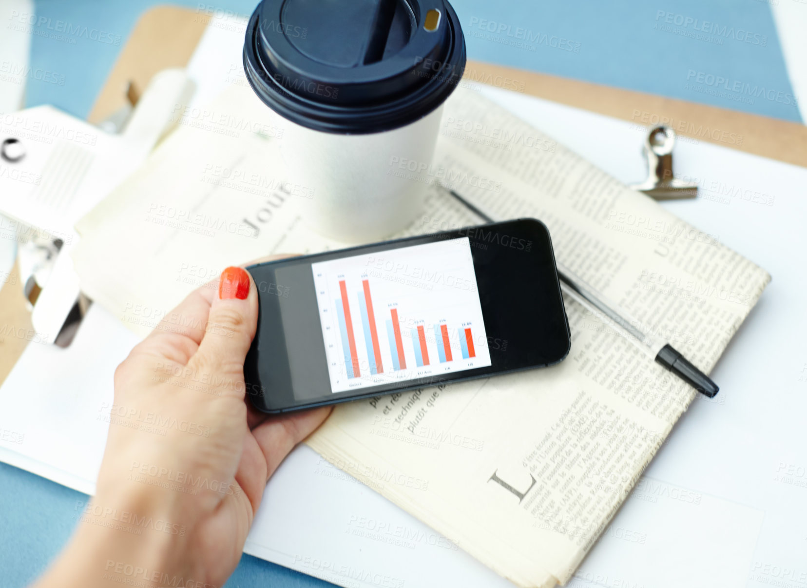 Buy stock photo Hand holding smartphone with bar chart on screen with disposable cup, pen, binder clip, newspaper and clipboard