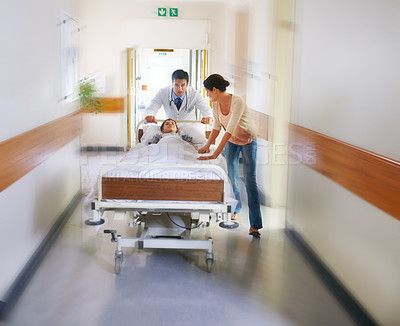 Buy stock photo Shot of a young boy being wheeled down a passage by his doctor and mother