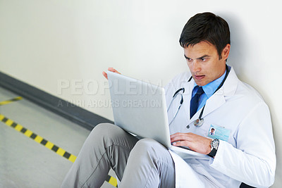 Buy stock photo Shot of a handsome young doctor sitting in a hospital corridor doing research on a laptop