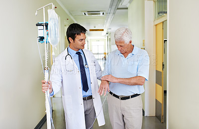 Buy stock photo Shot of a handsome young doctor walking with his senior patient down the hall