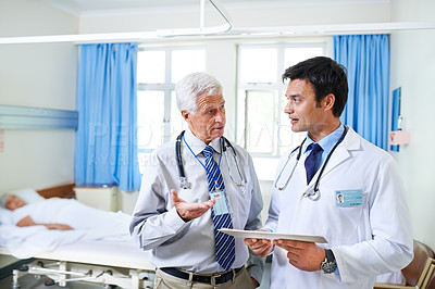 Buy stock photo Shot of two doctors discussing a case file with their patient in a hospital bed in the background