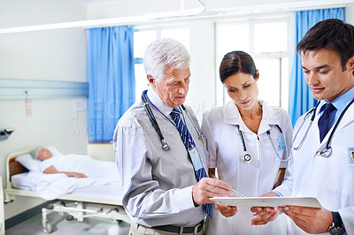 Buy stock photo Shot of three doctors discussing a case file with a patient in a hospital bed in the background
