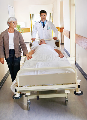 Buy stock photo Shot of a sick man on a bed being wheeled through a hospital corridor with his wife at his side