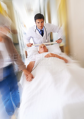 Buy stock photo Shot of a sick man on a bed being wheeled at speed through a hospital corridor with his wife at his side