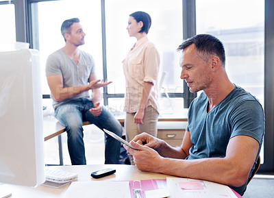 Buy stock photo Shot of an office worker using a tablet while his coworkers have a discussion in the background
