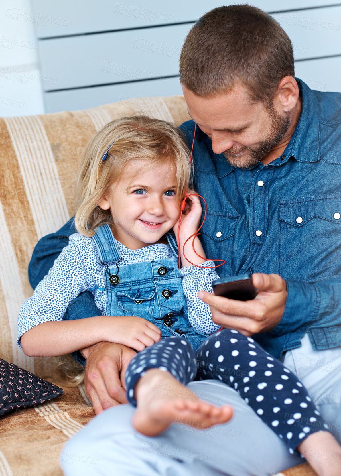 Buy stock photo Shot of an adorable little girl sitting on the sofa sharing headphones with her father