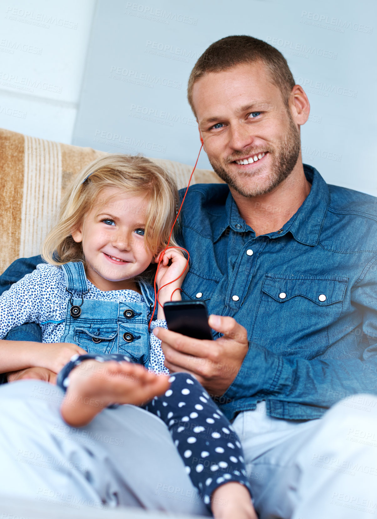 Buy stock photo Portrait of an adorable little girl sitting on the sofa sharing headphones with her father