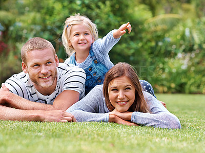 Buy stock photo Shot of a happy family lying outside on the grass with their young daughter pointing at something