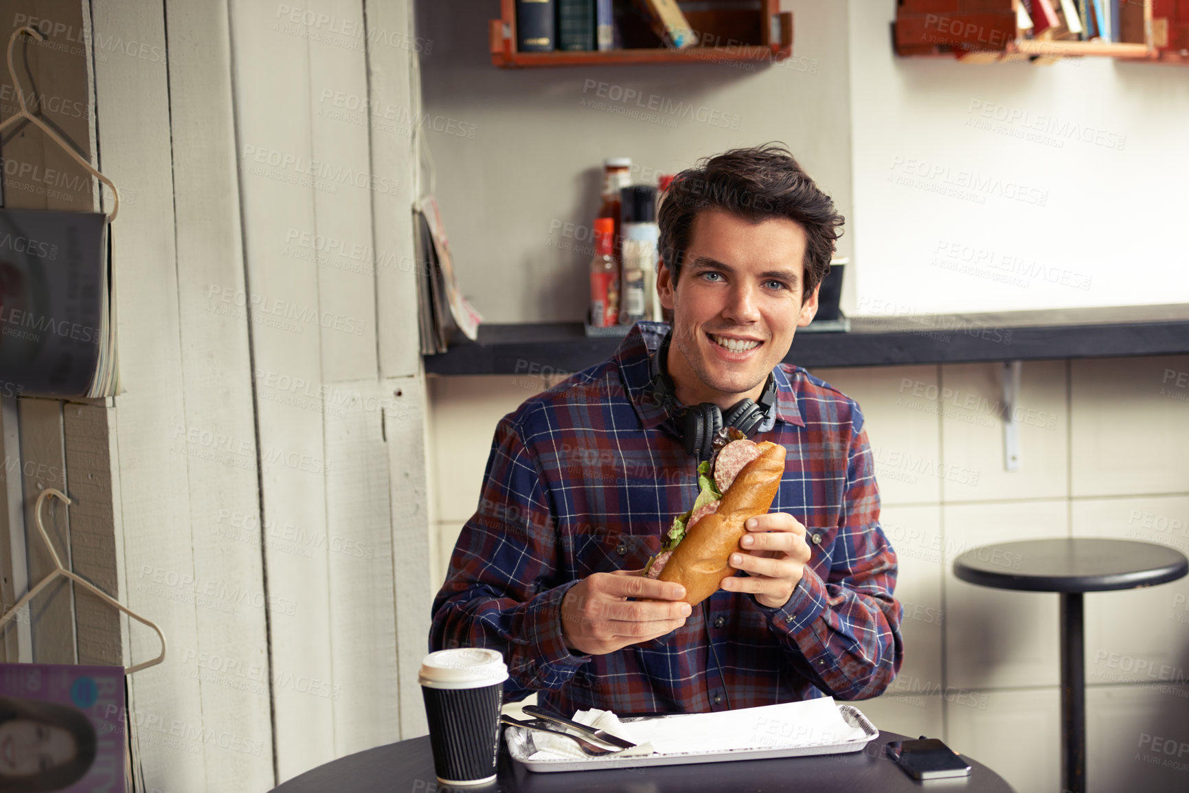 Buy stock photo Portrait of a hungry man eating a french baguette sandwich and a cup of coffee at a cafe while wearing wireless headphones. Happy customer enjoying a ham bread sandwich in a restaurant  alone