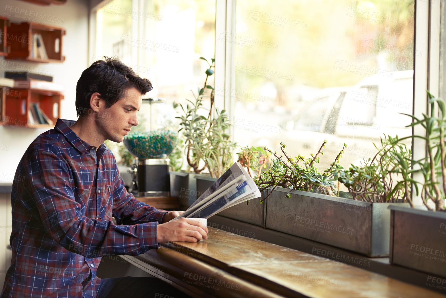 Buy stock photo Profile of a young male reading a newspaper and drinking coffee in the window of a modern cafe on a sunny day. Focused student or businessman alone checking the news. Old school current event updates