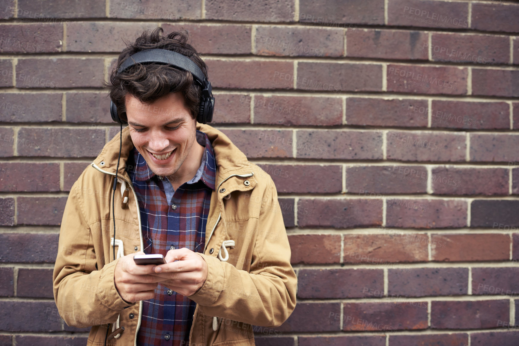 Buy stock photo Texting, headphones and happy man with phone by brick wall for communication, connection or chat. Smile, outdoor or student in city on urban street for music mobile app, social media or notification