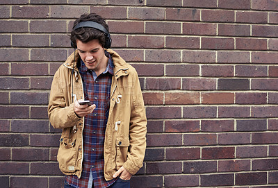 Buy stock photo Texting, headphones and man with mobile by brick wall for communication, connection or chat. Phone, outdoor or student in city on urban street for music app, social media website or news notification