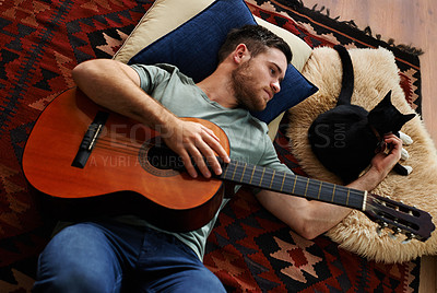 Buy stock photo Shot of a young man with a guitar lying on the floor with his cat
