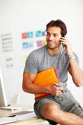 Buy stock photo A smiling handsome man sitting on an office desk and talking on his cellphone with folder under his arm and pen in hand