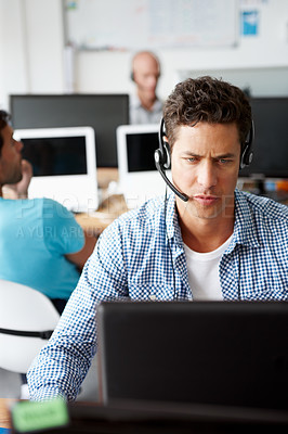 Buy stock photo A handsome young man sitting at a desk working on his computer and wearing a headset with a expression of concentration on his face