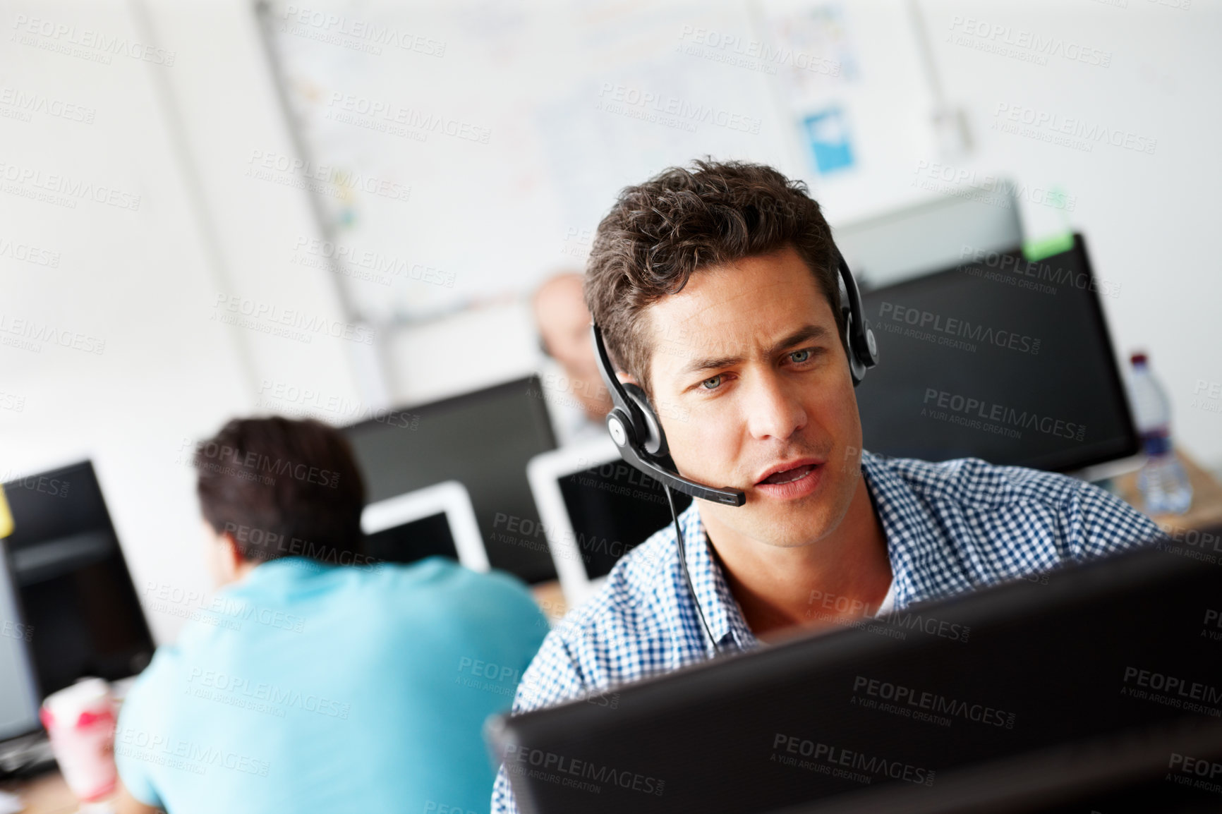 Buy stock photo A handsome young sales assistant talking on his headset while working on a computer and colleagues working in the background