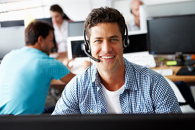 Buy stock photo Portrait of a handsome young sales assistant talking on his headset while working on a computer and colleagues working in the background