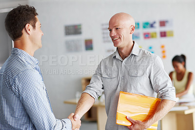 Buy stock photo Shot of two young men shaking hands in an office with a colleague working at a desk in the background