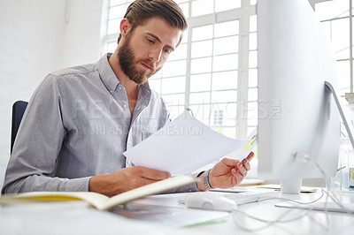 Buy stock photo Shot of a young designer holding paperwork while sitting in front of his computer