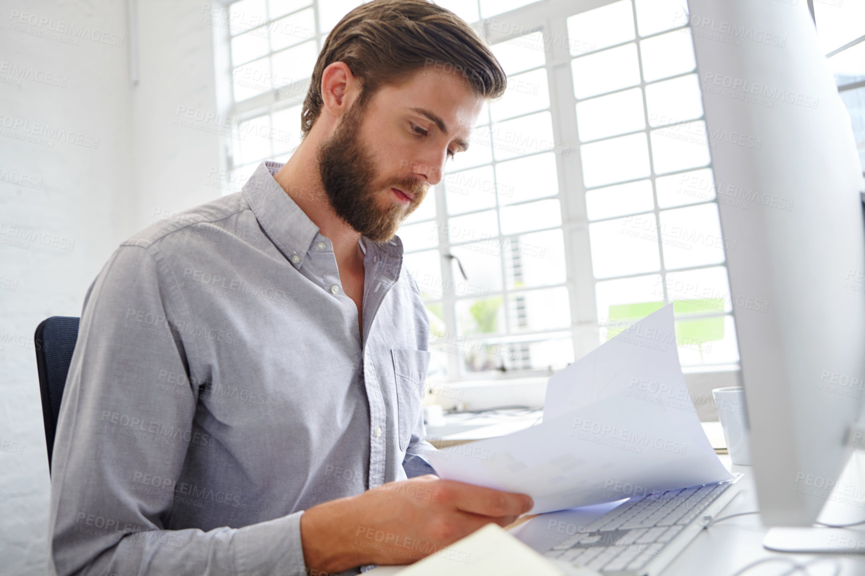 Buy stock photo Shot of a young designer holding paperwork while sitting in front of his computer