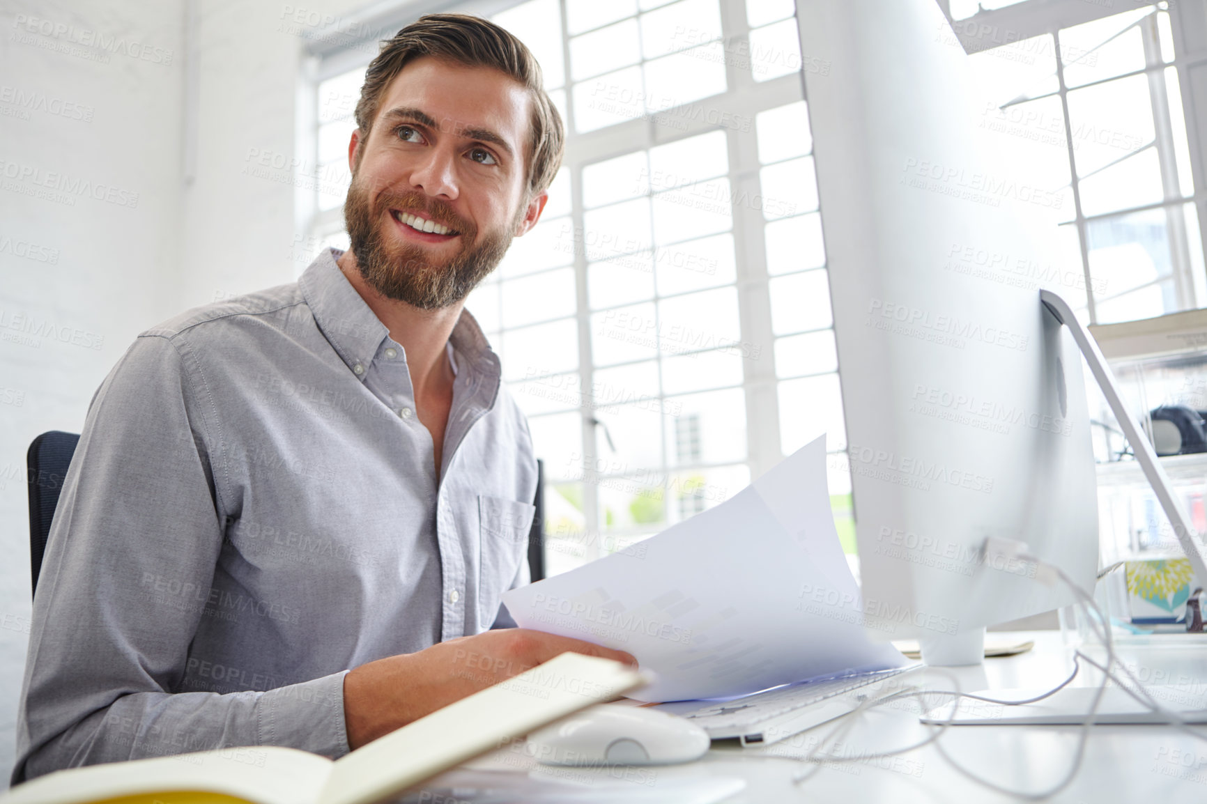 Buy stock photo Shot of a young designer holding paperwork while sitting in front of his computer