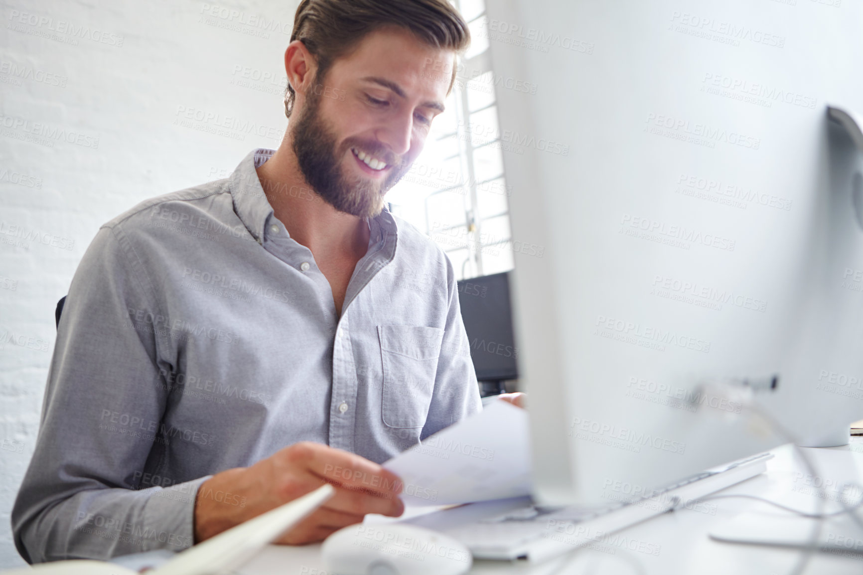 Buy stock photo Shot of a young designer holding paperwork while sitting in front of his computer