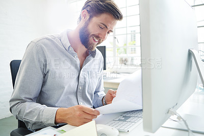 Buy stock photo Shot of a young designer holding paperwork while sitting in front of his computer