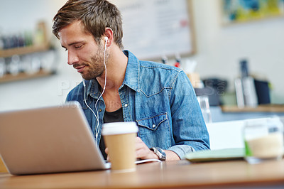 Buy stock photo Shot of a young man listening to music while using his laptop at a coffee shop