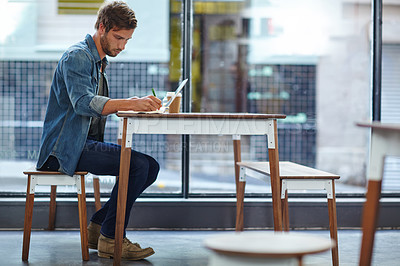 Buy stock photo Full length shot of a young man writing notes while sitting with his laptop at a coffee shop table