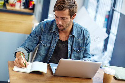 Buy stock photo Shot of a young man writing notes while sitting with his laptop at a coffee shop table