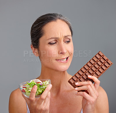 Buy stock photo Mature woman, salad bowl or chocolate with choice for diet, pick or selection in studio on a gray background. Female person, nutritionist or model with organic or mix vegetables and slab for decision