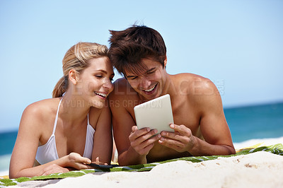 Buy stock photo Shot of a happy young couple watching something on their tablet at the beach