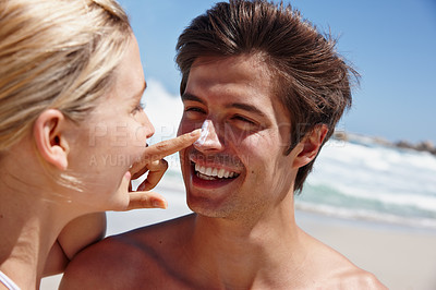 Buy stock photo Shot of a happy young woman applying sunscreen to her boyfriend's nose at the beach