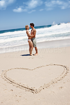 Buy stock photo Shot of a young couple enjoying a romantic moment at the beach next to a drawing of a heart in the sand