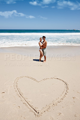 Buy stock photo Shot of a young couple enjoying a romantic moment at the beach next to a drawing of a heart in the sand