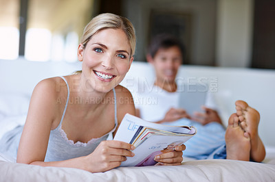 Buy stock photo Shot of a happy young woman lying in bed and reading a magazine while her husband uses a digital tablet in the background