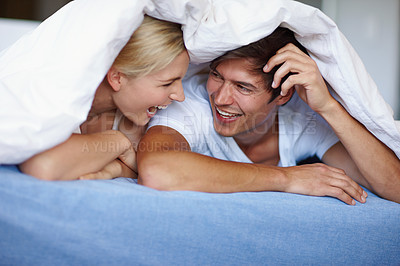 Buy stock photo Shot of a happy young couple enjoying a playful moment underneath the duvet