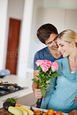 Buy stock photo Shot of a handsome young man giving his wife a bouquet of pink roses while she prepares a meal