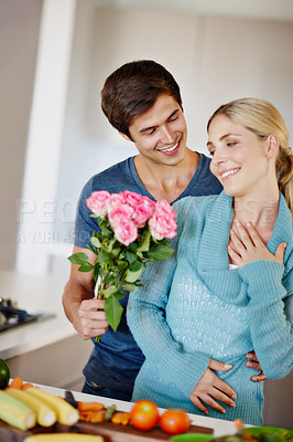 Buy stock photo Shot of a handsome young man giving his wife a bouquet of pink roses while she prepares a meal