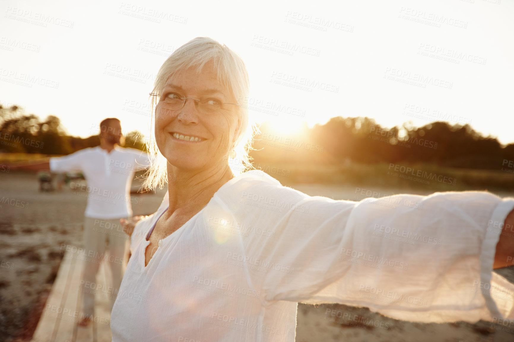 Buy stock photo Shot of a mature woman doing yoga on the beach with her husband in the background