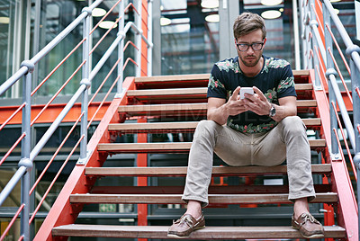 Buy stock photo Shot of a handsome young man using his mobile phone while sitting on the steps of an office 