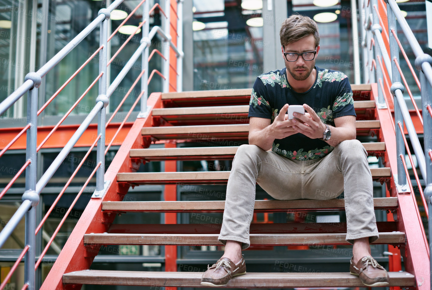 Buy stock photo Shot of a handsome young man using his mobile phone while sitting on the steps of an office 