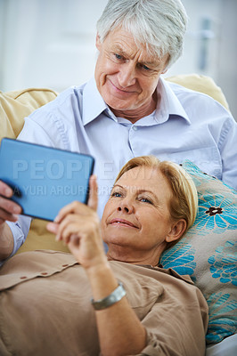 Buy stock photo Shot of a happy elderly couple watching something on a digital tablet while relaxing on their sofa