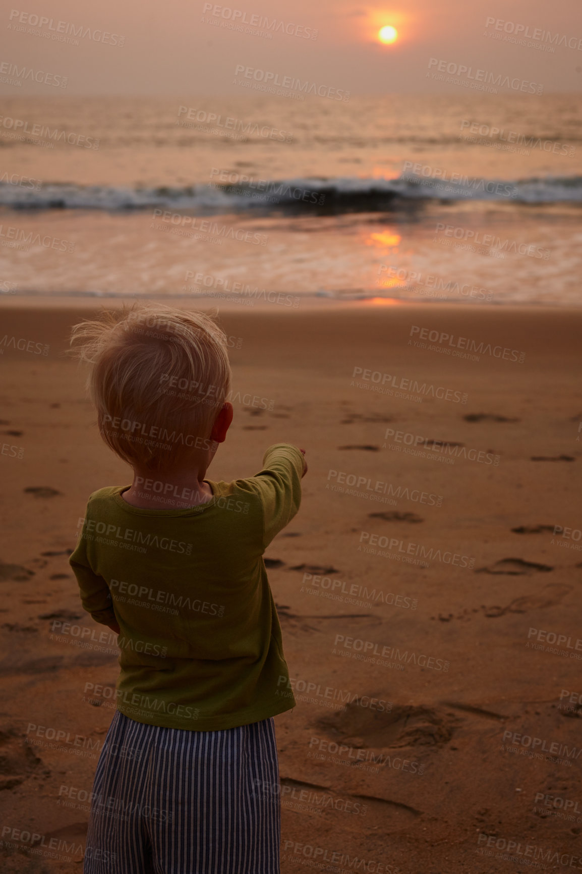 Buy stock photo Rearview of a little boy on a beach at sunset pointing to the setting sun