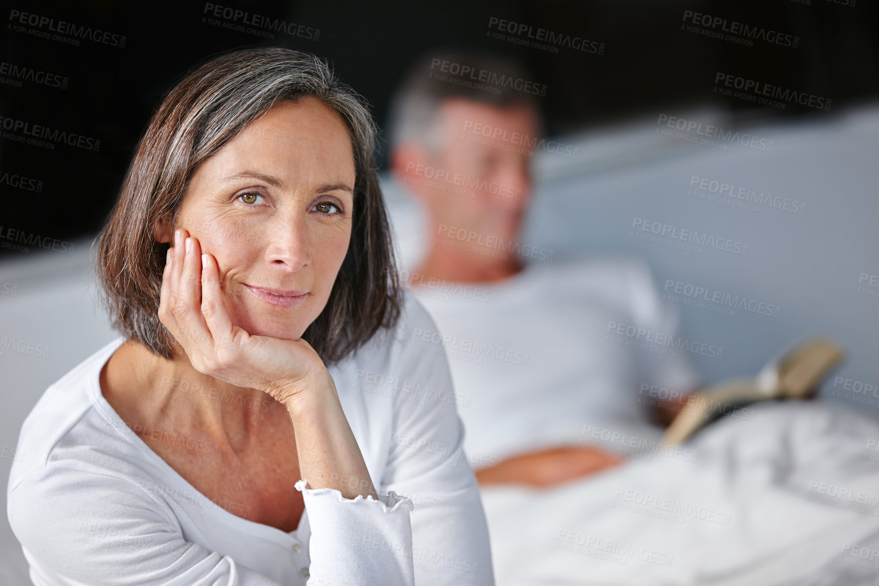 Buy stock photo Portrait of an attractive mature woman sitting on her bed with her husband in the background