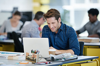 Buy stock photo Shot of a designer sitting at his desk working on a laptop with colleagues in the background