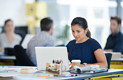 Buy stock photo Shot of a designer sitting at her desk working on a laptop with colleagues in the background