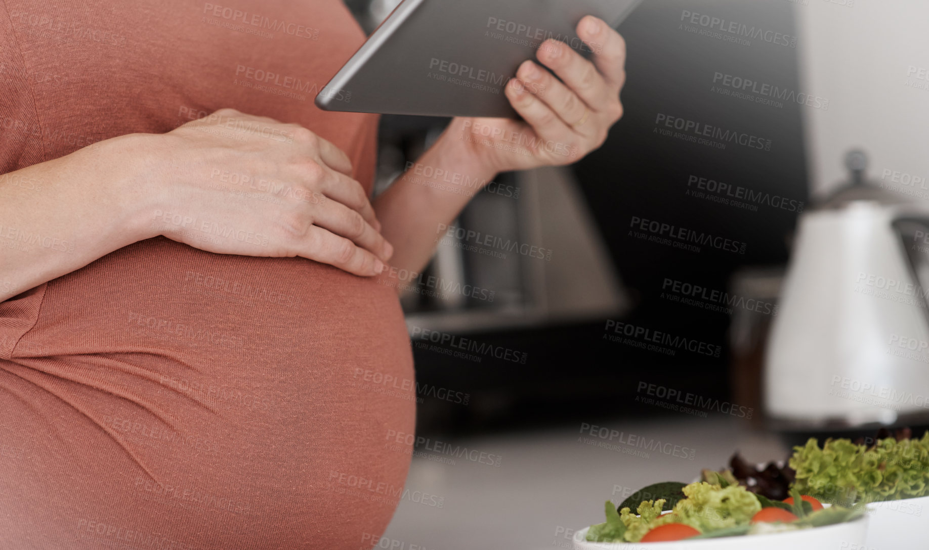 Buy stock photo Cropped shot of a pregnant woman using her tablet while eating a salad in the kitchen