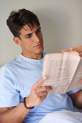 Buy stock photo Shot of a handsome young man reading a newspaper in his bedroom